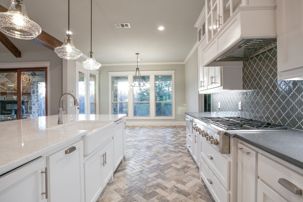Design ideas for a large traditional l-shaped open plan kitchen in Dallas with a farmhouse sink, shaker cabinets, white cabinets, granite benchtops, grey splashback, glass tile splashback, stainless steel appliances, brick floors, with island and grey floor.
