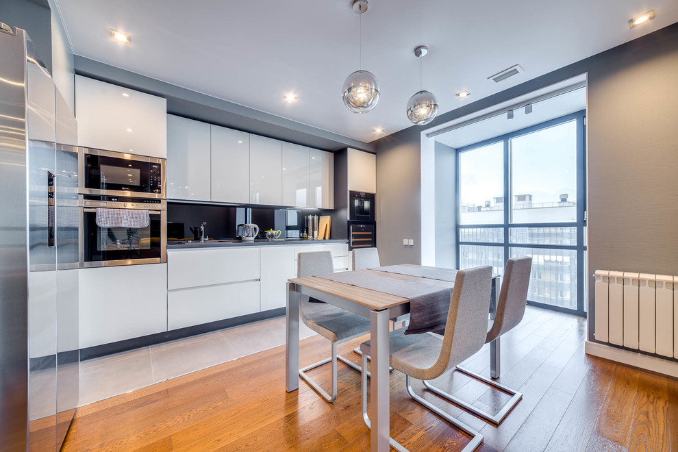 Contemporary l-shaped eat-in kitchen in Saint Petersburg with flat-panel cabinets, white cabinets, black splashback, glass sheet splashback, stainless steel appliances, beige floor and grey benchtop.