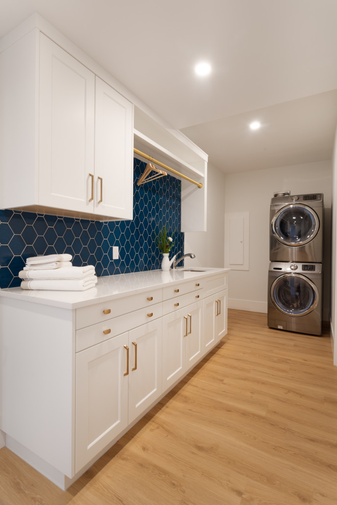 This is an example of a mid-sized transitional galley dedicated laundry room in Calgary with a single-bowl sink, shaker cabinets, white cabinets, quartz benchtops, blue splashback, ceramic splashback, white walls, light hardwood floors, a stacked washer and dryer, beige floor and white benchtop.