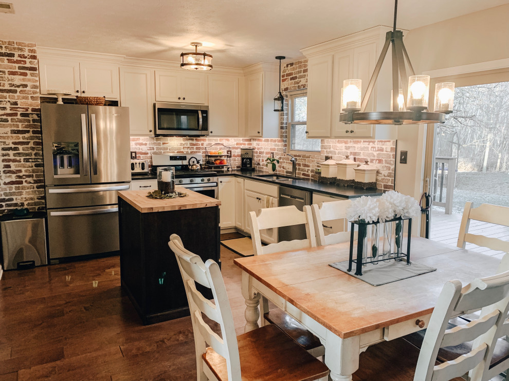 This is an example of a small country l-shaped eat-in kitchen in Indianapolis with an undermount sink, recessed-panel cabinets, white cabinets, granite benchtops, brown splashback, brick splashback, stainless steel appliances, medium hardwood floors, with island, brown floor and black benchtop.