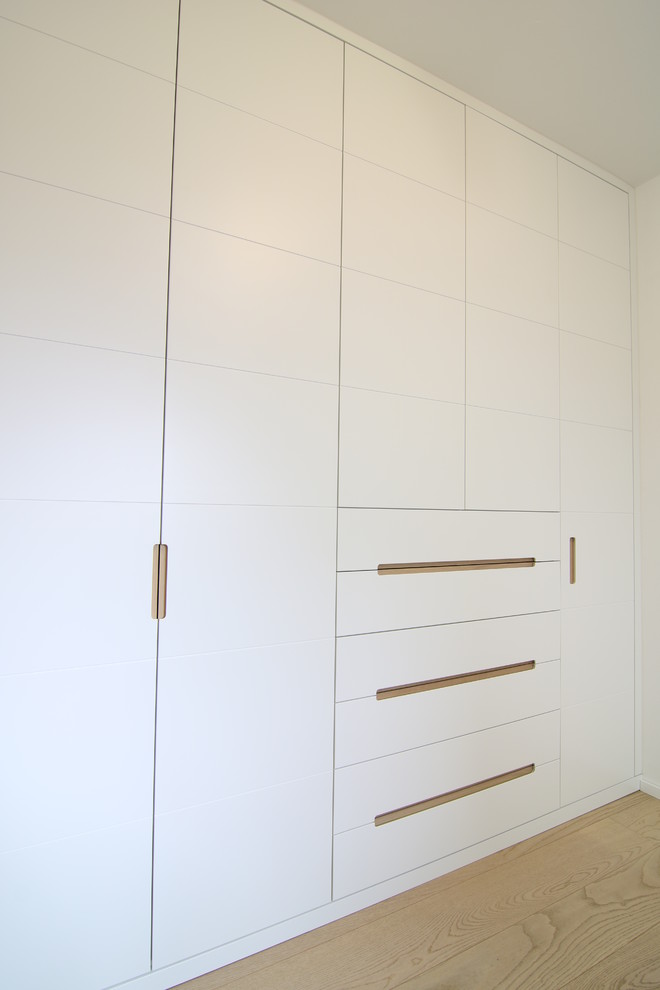 Diseño de armario unisex actual de tamaño medio con armarios con paneles lisos, puertas de armario blancas y suelo de madera clara