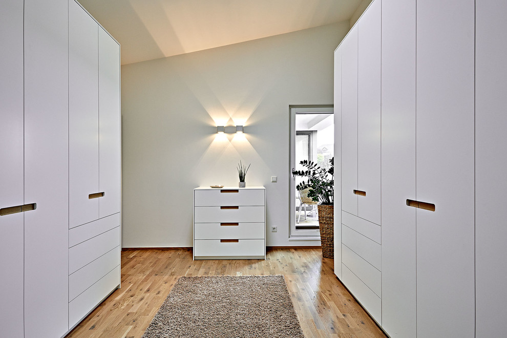 Modelo de armario vestidor unisex contemporáneo de tamaño medio con armarios con paneles lisos, puertas de armario blancas, suelo de madera en tonos medios y suelo marrón