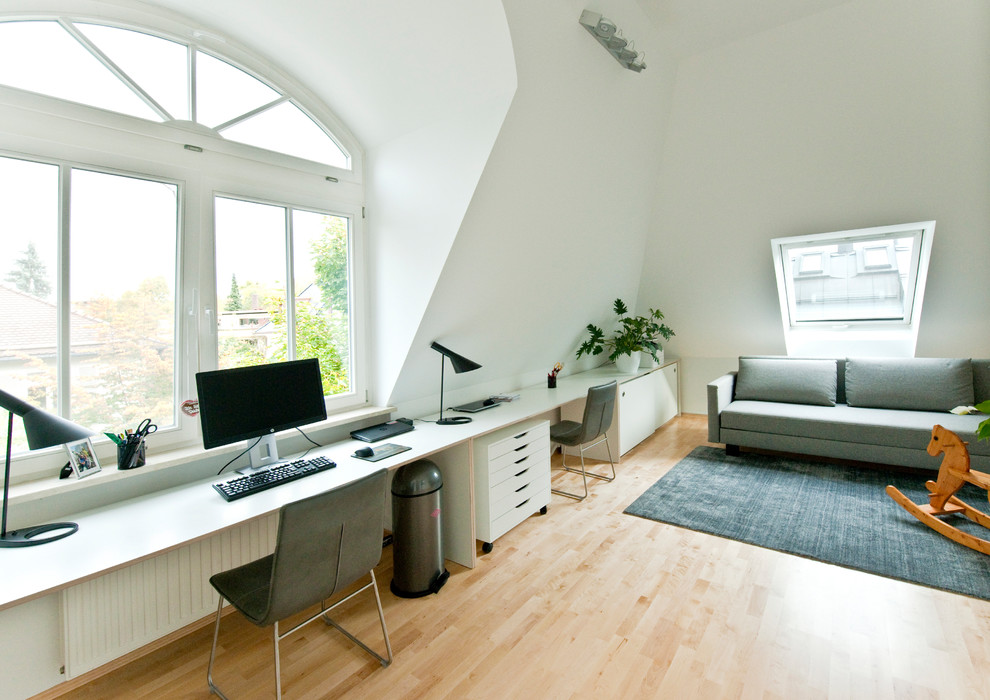 Modelo de despacho contemporáneo grande sin chimenea con paredes blancas, suelo de madera clara, escritorio empotrado y suelo marrón