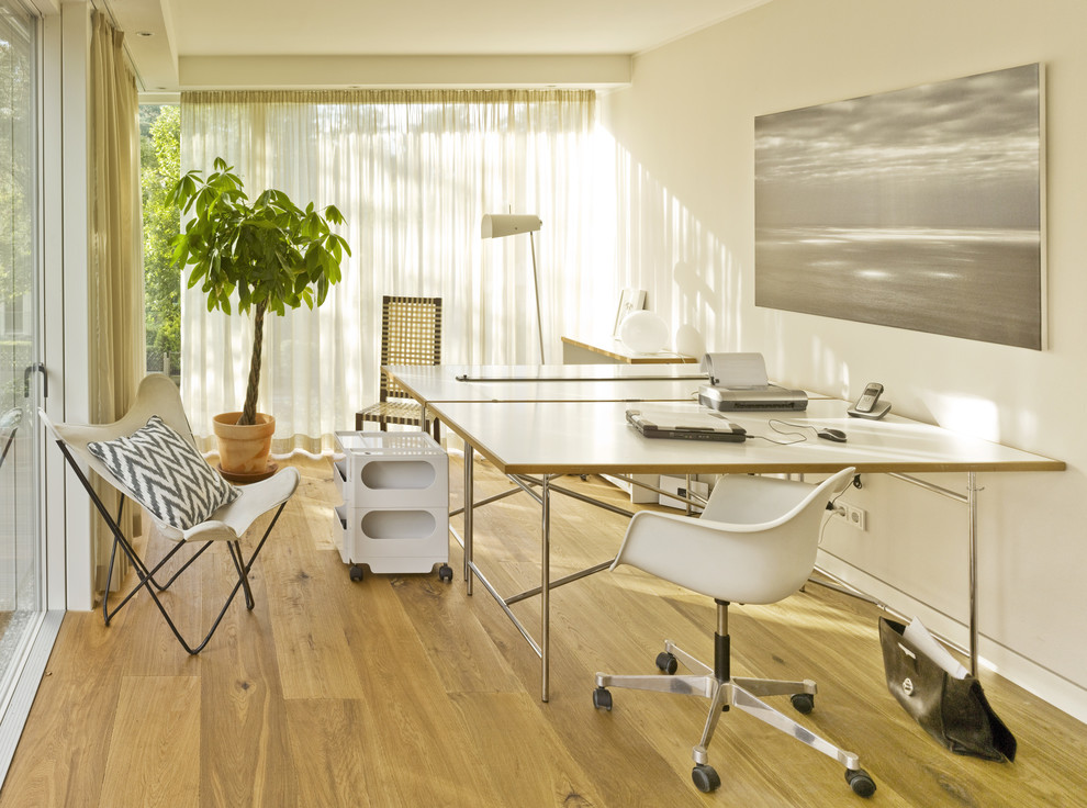 Пример оригинального дизайна: большое рабочее место в современном стиле с белыми стенами, светлым паркетным полом и отдельно стоящим рабочим столом без камина