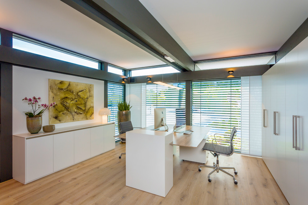 Diseño de despacho actual grande sin chimenea con paredes blancas, suelo de madera clara, escritorio independiente y suelo beige