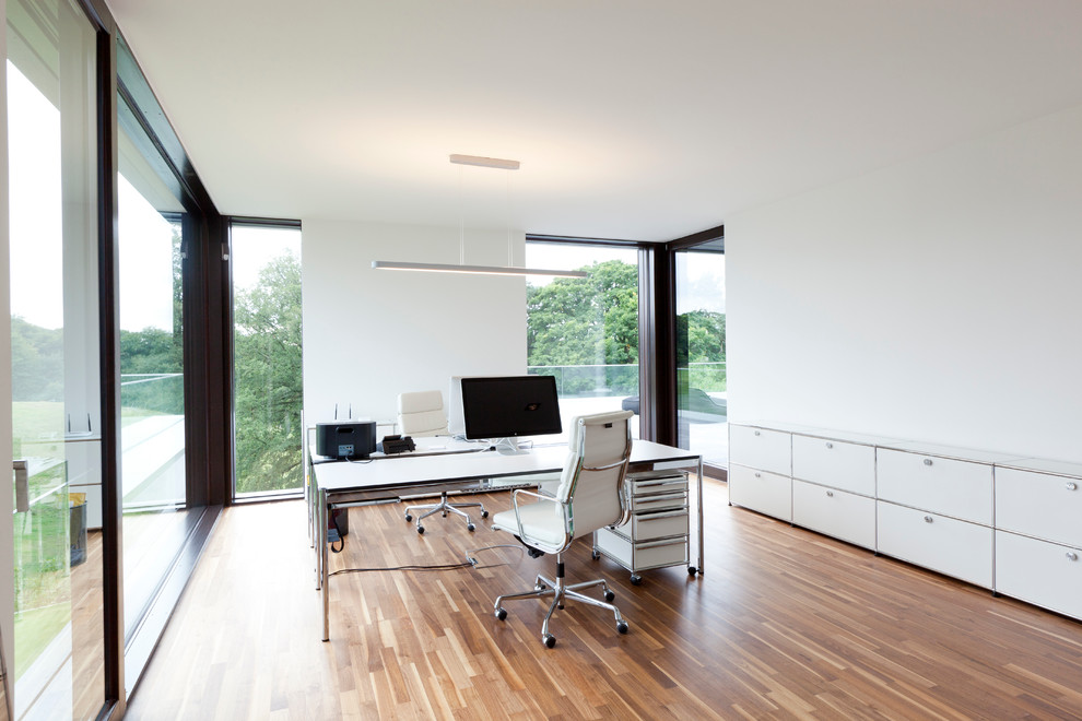 Пример оригинального дизайна: большое рабочее место в стиле модернизм с белыми стенами, паркетным полом среднего тона и отдельно стоящим рабочим столом без камина