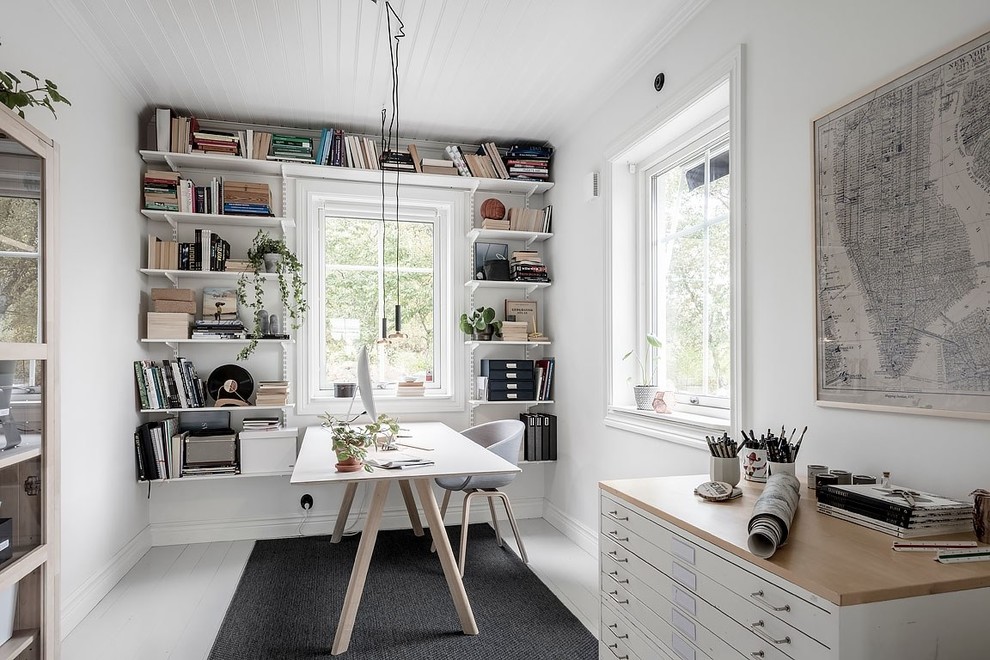 На фото: домашняя библиотека в скандинавском стиле с белыми стенами, деревянным полом, отдельно стоящим рабочим столом и белым полом без камина