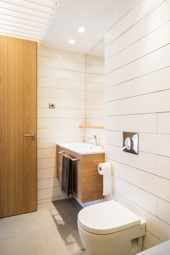 Minimalistisk inredning av ett litet toalett, med en vägghängd toalettstol, vit kakel, vita väggar, cementgolv och ett integrerad handfat