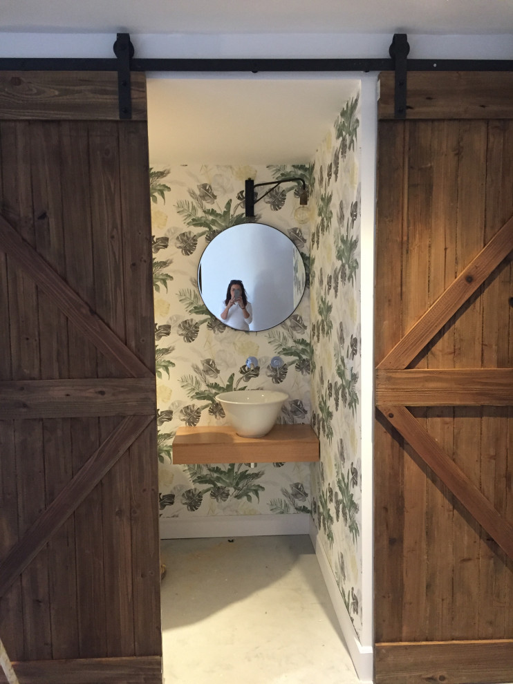 Cette photo montre un petit WC et toilettes montagne avec des portes de placard blanches, sol en béton ciré, une vasque, un sol gris, un plan de toilette marron, du papier peint, un plan de toilette en bois et meuble-lavabo suspendu.