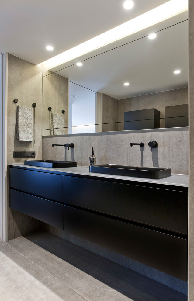 Example of a mid-sized trendy gray tile and ceramic tile ceramic tile and gray floor powder room design in Other with flat-panel cabinets, black cabinets, a wall-mount toilet, gray walls, a vessel sink, glass countertops and gray countertops