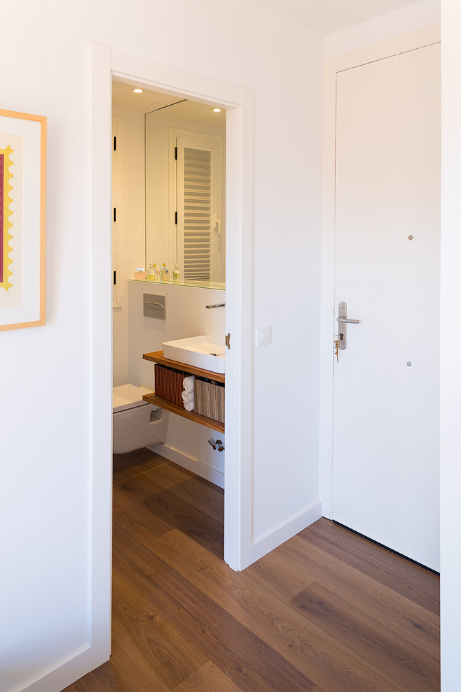 This is an example of a small contemporary cloakroom in Barcelona with open cabinets, medium wood cabinets, a wall mounted toilet, white tiles, cement tiles, white walls, medium hardwood flooring, a vessel sink, wooden worktops, brown floors and brown worktops.
