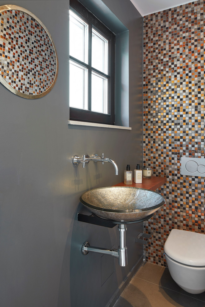 Modern inredning av ett brun brunt toalett, med flerfärgad kakel, mosaik, grå väggar, ett fristående handfat, träbänkskiva och en vägghängd toalettstol