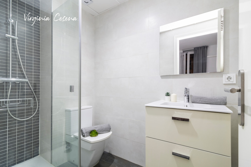 Skandinavisk inredning av ett mellanstort vit vitt badrum, med möbel-liknande, grå skåp, en toalettstol med hel cisternkåpa, vit kakel, keramikplattor, vita väggar, klinkergolv i porslin, ett väggmonterat handfat och grått golv