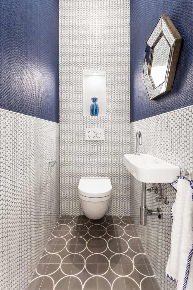 Bild på ett litet funkis toalett, med en vägghängd toalettstol, blå kakel, vit kakel, mosaik, blå väggar, ett väggmonterat handfat och grått golv