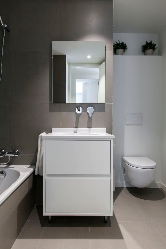 Bild på ett litet minimalistiskt toalett, med släta luckor, en vägghängd toalettstol, grå kakel, porslinskakel, grå väggar, klinkergolv i porslin, ett undermonterad handfat, bänkskiva i kvarts och vita skåp