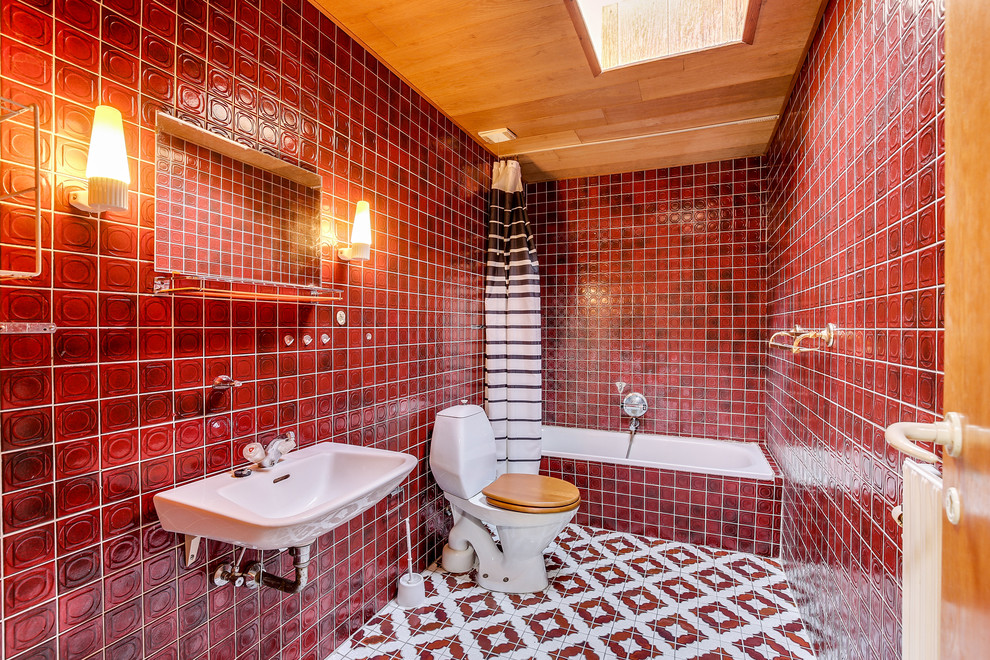 Bild på ett 60 tals badrum, med ett platsbyggt badkar, en toalettstol med separat cisternkåpa, röd kakel, röda väggar, ett väggmonterat handfat, flerfärgat golv och dusch med duschdraperi