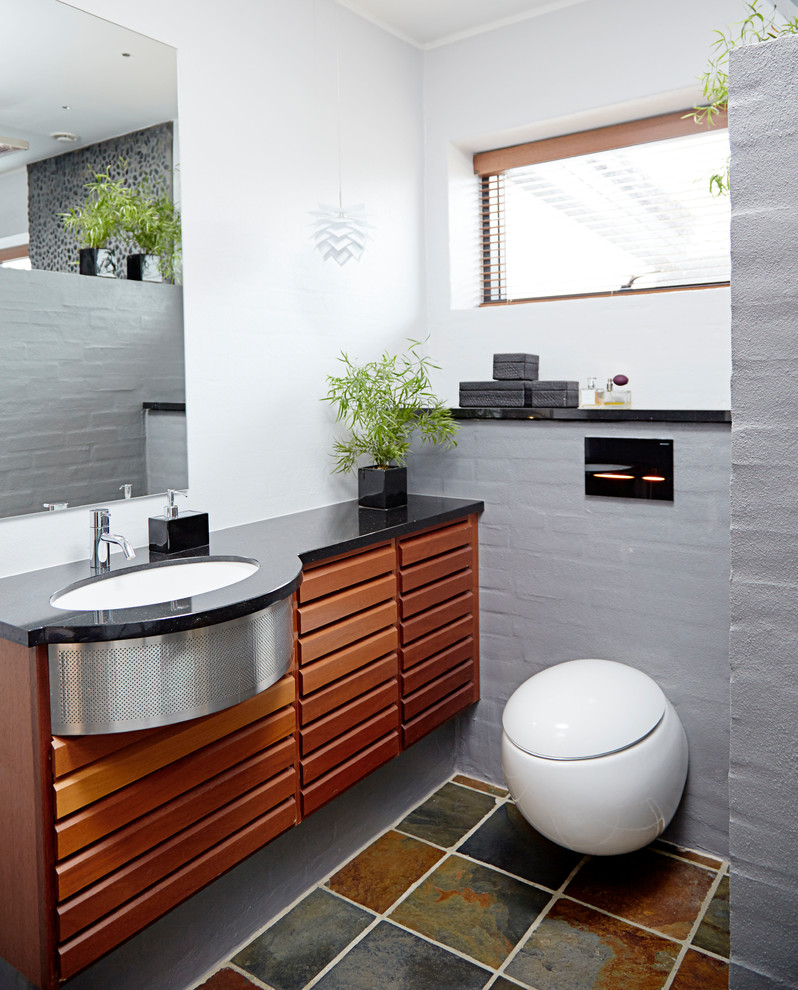 Cette image montre une salle de bain principale design en bois brun avec WC suspendus, un carrelage gris, une plaque de galets, un mur blanc, un lavabo encastré, un sol multicolore et un plan de toilette noir.