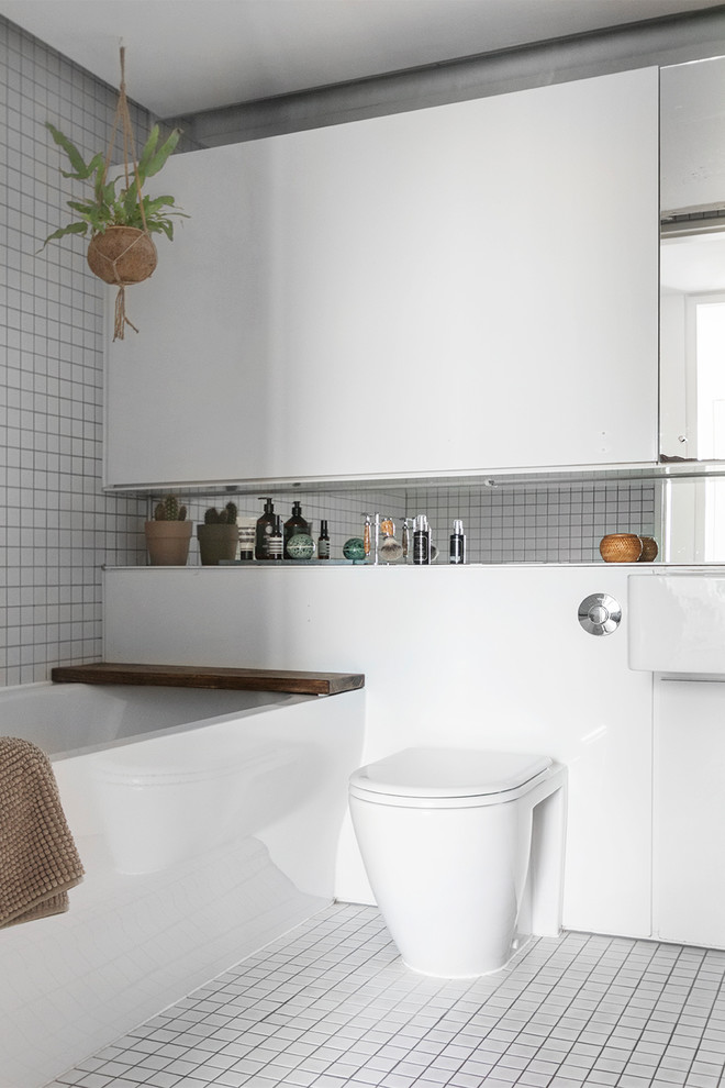 This is an example of a scandinavian shower room bathroom in Aarhus with flat-panel cabinets, white cabinets, a corner bath, a wall mounted toilet, white tiles, mosaic tiles, white walls, mosaic tile flooring, a wall-mounted sink and white floors.