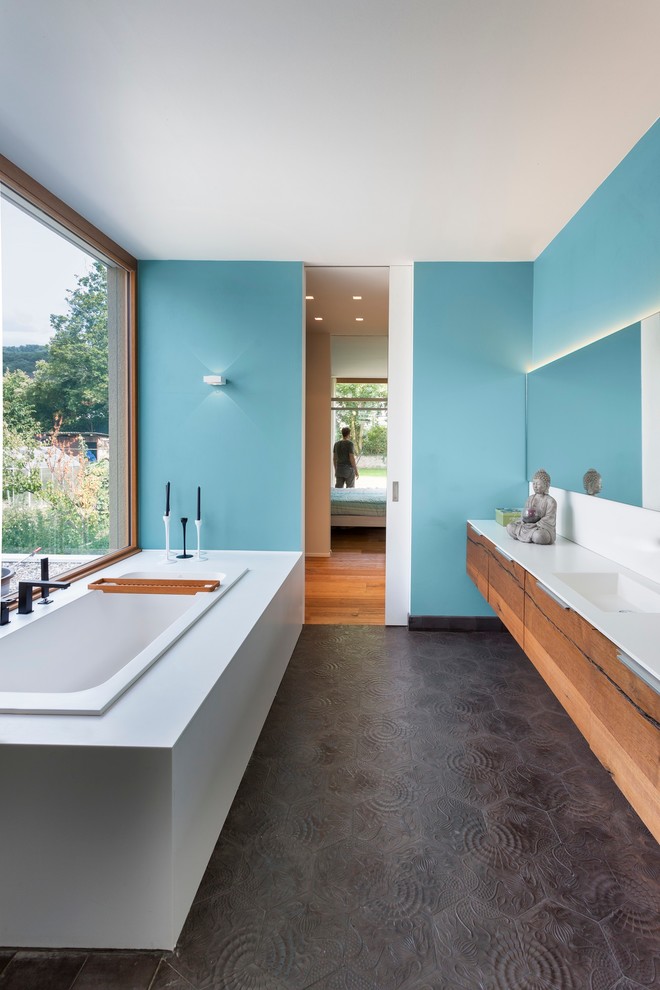 Cette image montre une grande salle de bain principale design en bois brun avec un lavabo intégré, un placard à porte plane, une baignoire posée, un mur bleu, un carrelage noir, des carreaux de béton et un plan de toilette en surface solide.