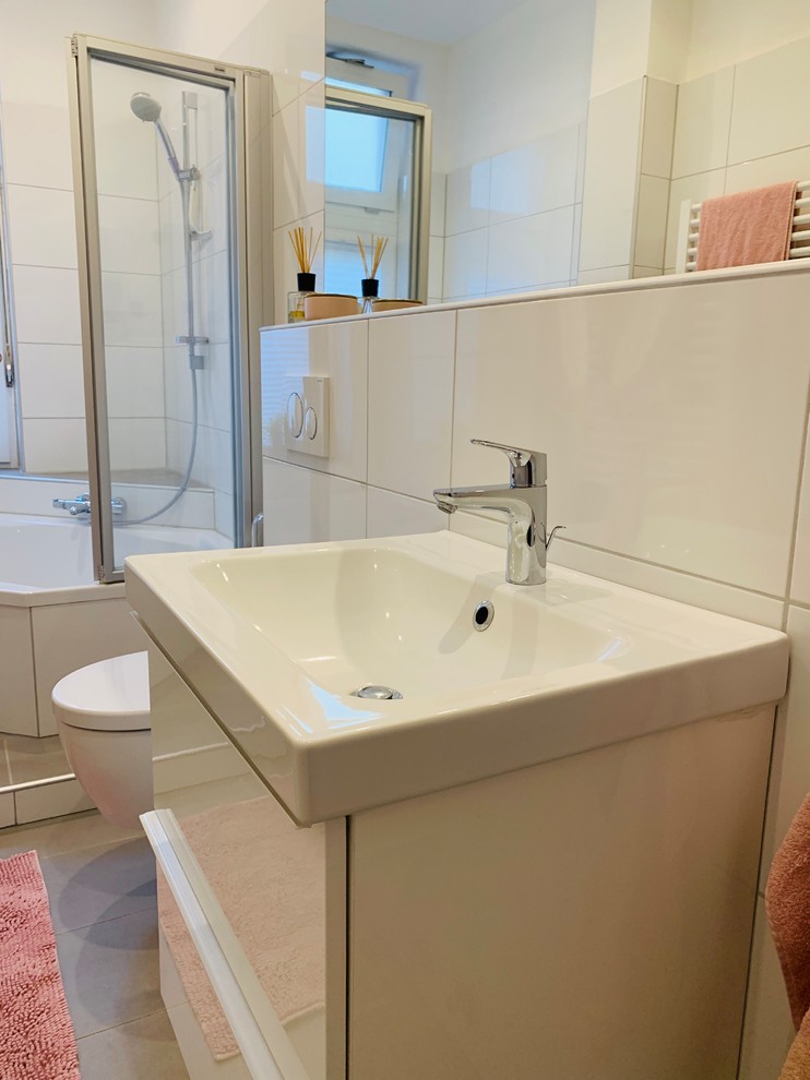 This is an example of a medium sized contemporary shower room bathroom in Hanover with white cabinets, a corner bath, a shower/bath combination, a wall mounted toilet, white tiles, ceramic tiles, white walls, ceramic flooring, a wall-mounted sink, solid surface worktops, grey floors, a hinged door and white worktops.