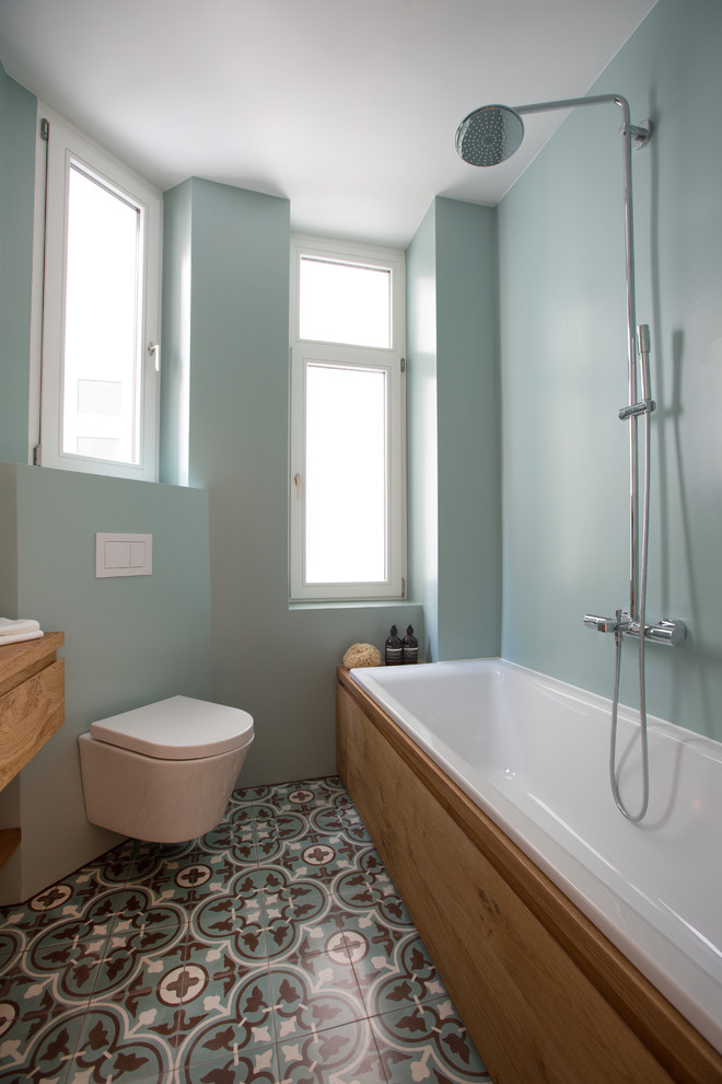 This is an example of a small scandinavian bathroom in Frankfurt with flat-panel cabinets, medium wood cabinets, a built-in bath, a shower/bath combination, a wall mounted toilet, green walls, cement flooring, a vessel sink, wooden worktops, multi-coloured floors and a shower curtain.