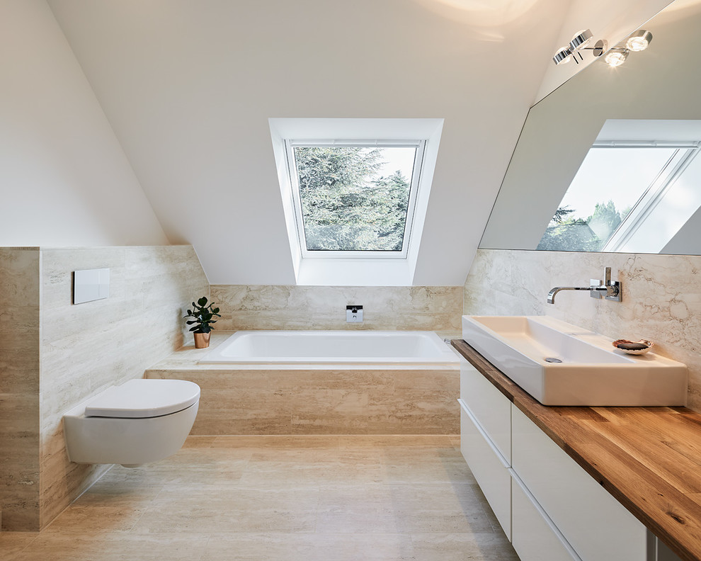 Small trendy travertine tile travertine floor drop-in bathtub photo in Dusseldorf with flat-panel cabinets, white cabinets, a wall-mount toilet, white walls, a vessel sink, wood countertops and brown countertops