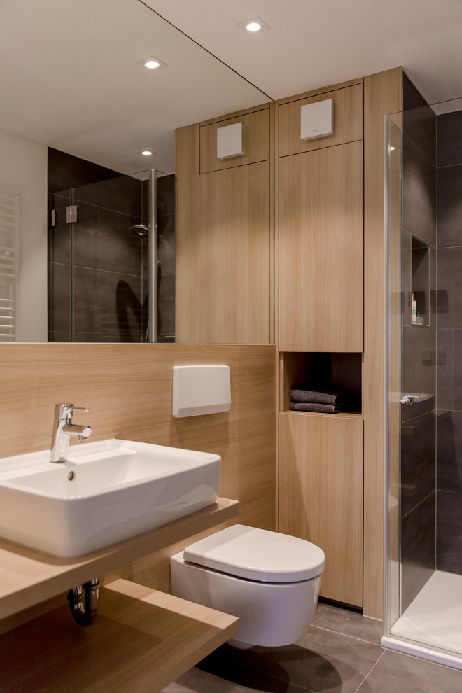 Inspiration for a contemporary shower room bathroom with flat-panel cabinets, light wood cabinets, a built-in shower, a wall mounted toilet, grey tiles, brown walls, a vessel sink, wooden worktops, grey floors, an open shower and brown worktops.