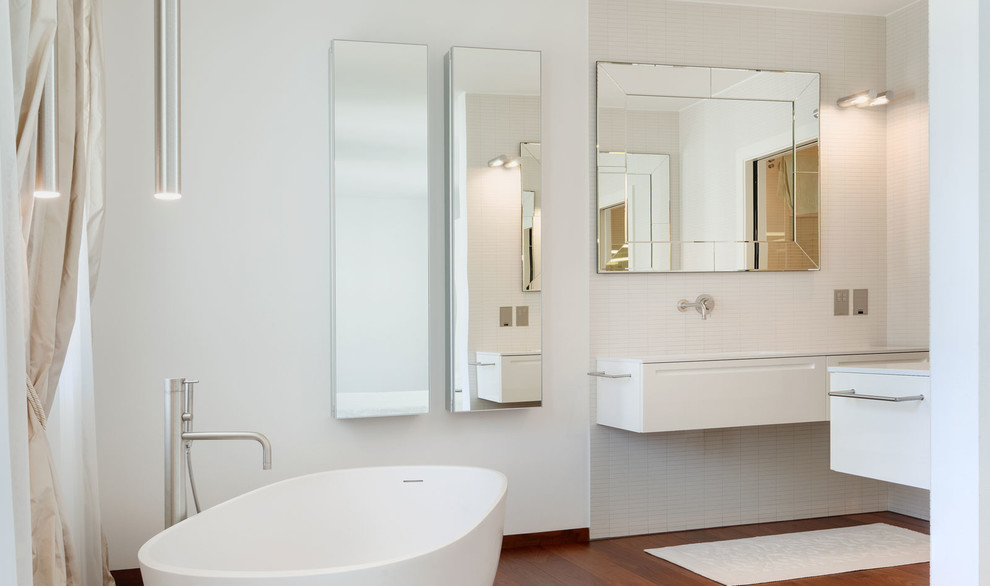 Bild på ett funkis badrum, med vita skåp, vit kakel, ett fristående badkar, keramikplattor, vita väggar, mörkt trägolv och ett integrerad handfat