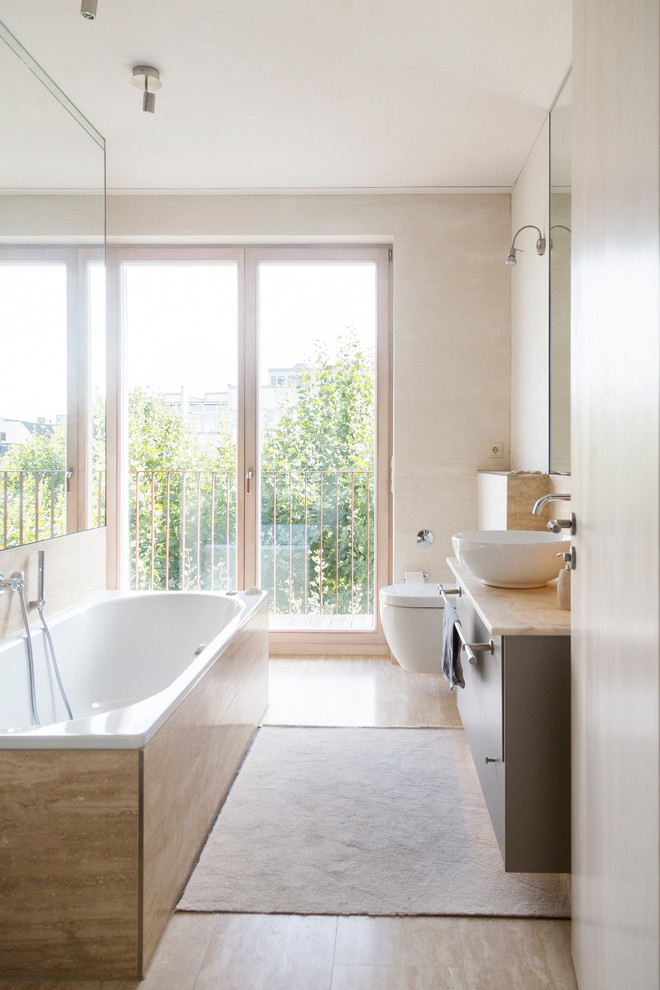 Inredning av ett modernt badrum, med ett fristående handfat, släta luckor, bruna skåp, ett platsbyggt badkar, beige väggar, en vägghängd toalettstol och marmorbänkskiva