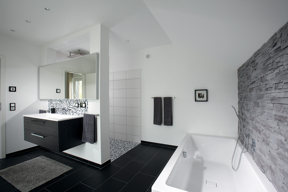 Modernes Badezimmer mit schwarzen Schränken, Einbaubadewanne, bodengleicher Dusche, grauen Fliesen, Steinplatten, weißer Wandfarbe, Einbauwaschbecken, schwarzem Boden, offener Dusche, weißer Waschtischplatte und Steinwänden in Sonstige