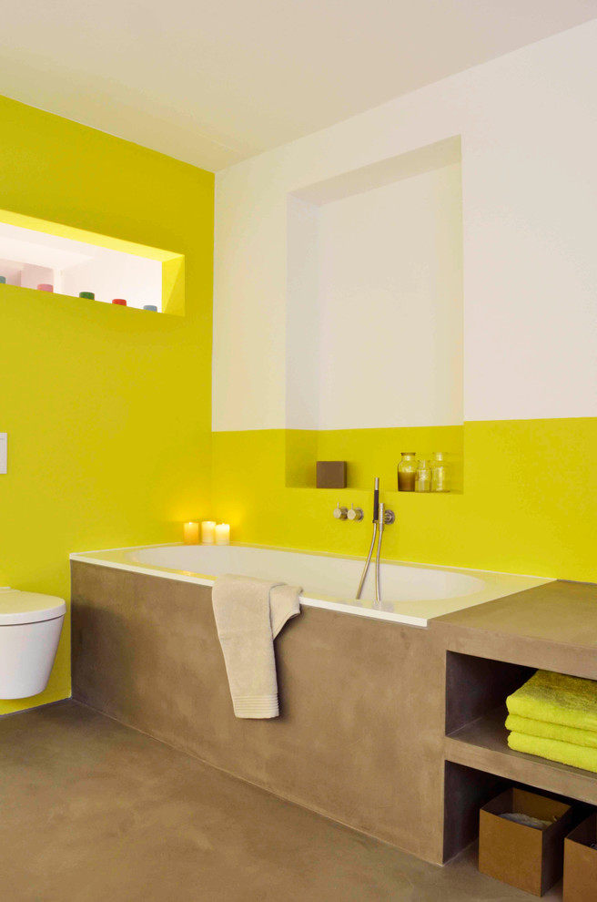 Réalisation d'une salle de bain design avec un placard sans porte, des portes de placard marrons, une baignoire posée, WC suspendus et un mur jaune.