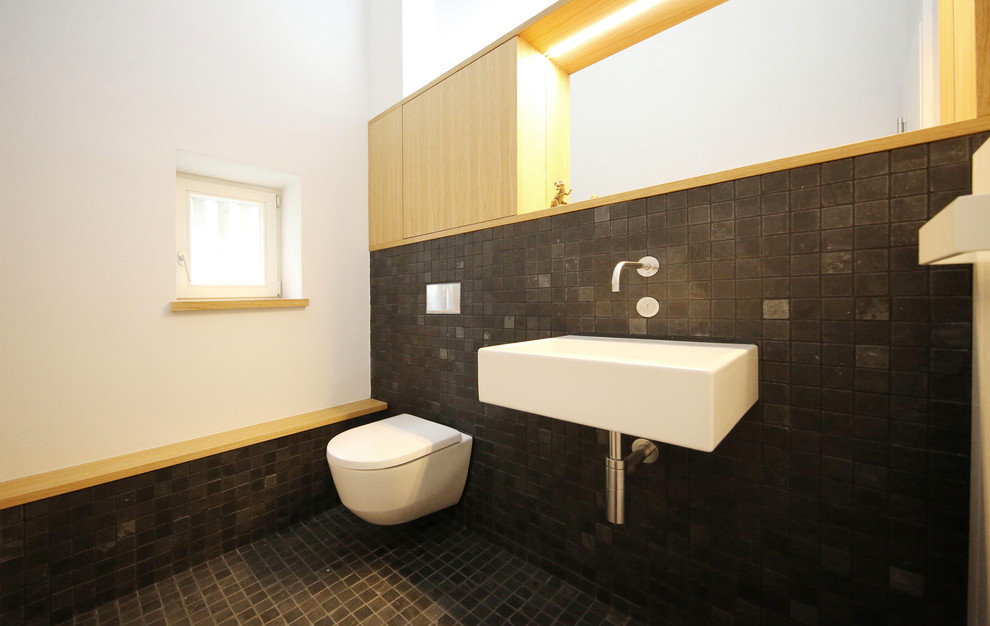 Bild på ett mellanstort funkis badrum med dusch, med släta luckor, skåp i ljust trä, en vägghängd toalettstol, svart kakel, vita väggar, ett väggmonterat handfat, mosaik och mosaikgolv