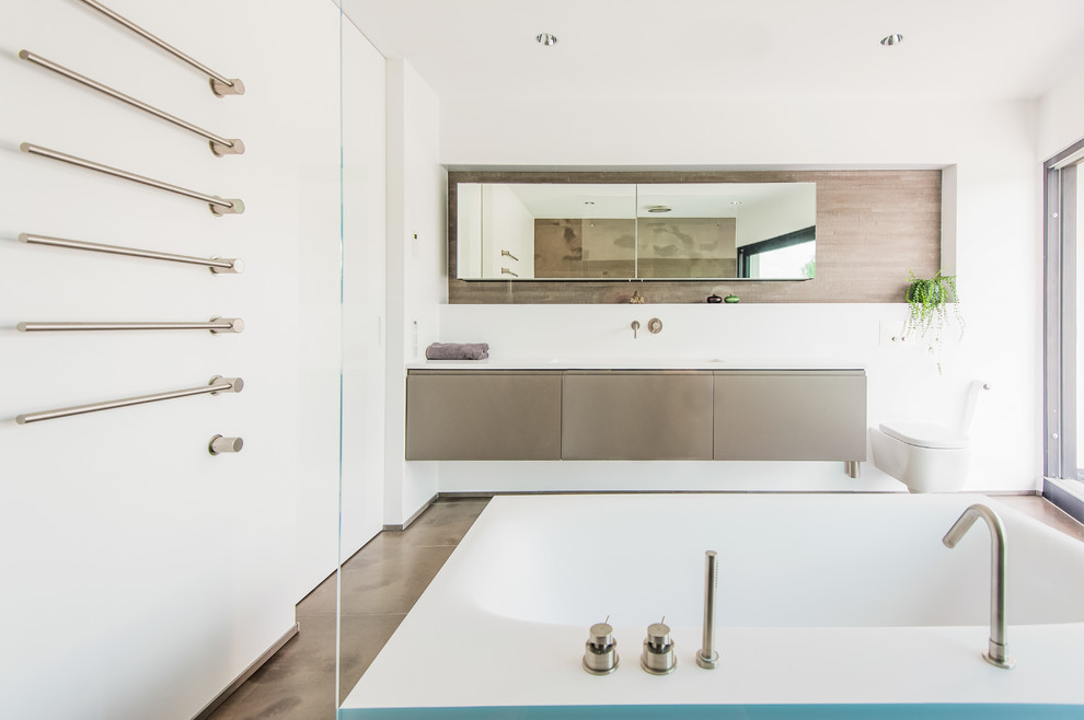 This is an example of a large contemporary bathroom in Frankfurt with flat-panel cabinets, grey cabinets, a wall mounted toilet, white walls, an integrated sink, solid surface worktops, a freestanding bath, brown tiles and stone tiles.
