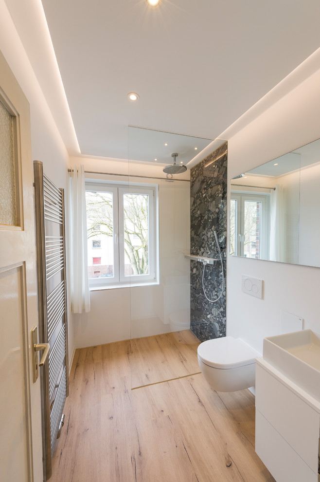 Bild på ett mellanstort funkis vit vitt badrum med dusch, med vita skåp, en kantlös dusch, en vägghängd toalettstol, vita väggar, mellanmörkt trägolv, ett fristående handfat, brunt golv, med dusch som är öppen, släta luckor, svart kakel, kakel i småsten och bänkskiva i akrylsten