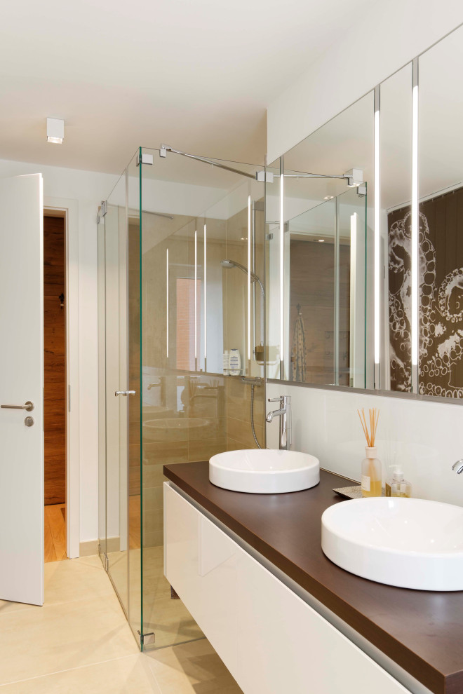Modernes Duschbad mit weißen Schränken, bodengleicher Dusche, Wandtoilette, beigen Fliesen, Keramikfliesen, weißer Wandfarbe, Keramikboden, Aufsatzwaschbecken, Waschtisch aus Holz, beigem Boden und brauner Waschtischplatte in Sonstige