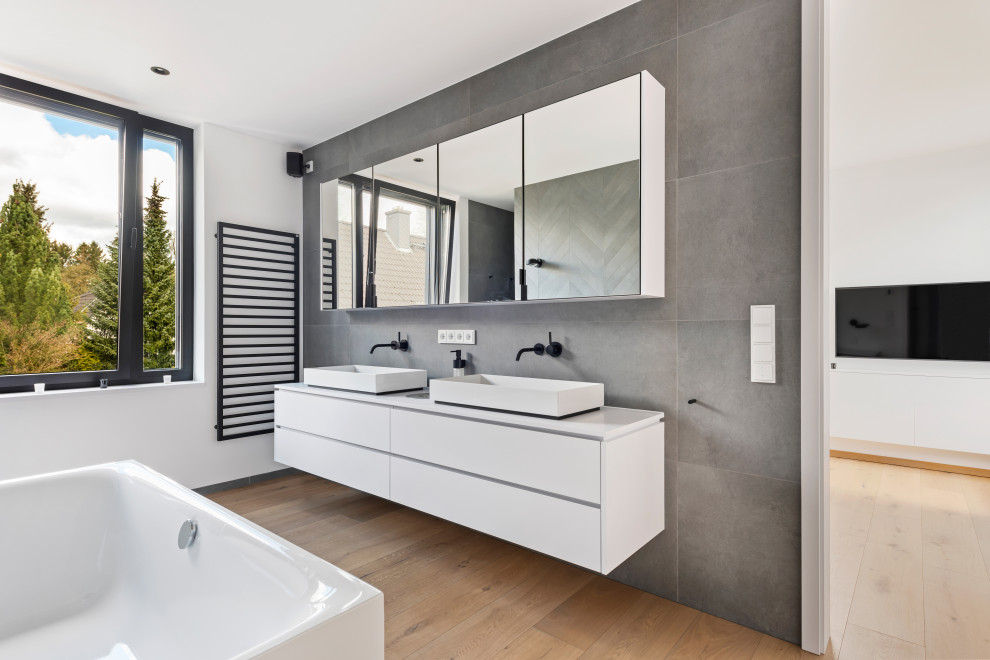 Exemple d'une salle de bain tendance de taille moyenne avec un placard à porte plane, des portes de placard blanches, une baignoire indépendante, un carrelage gris, un sol en bois brun, une vasque, un sol marron, un plan de toilette blanc, meuble double vasque et meuble-lavabo suspendu.