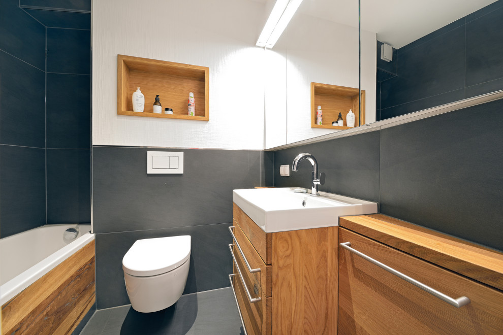 Inredning av ett modernt litet badrum med dusch, med luckor med infälld panel, skåp i mellenmörkt trä, ett platsbyggt badkar, en toalettstol med separat cisternkåpa, svart kakel, stenkakel, vita väggar, terrazzogolv, ett fristående handfat, träbänkskiva och svart golv