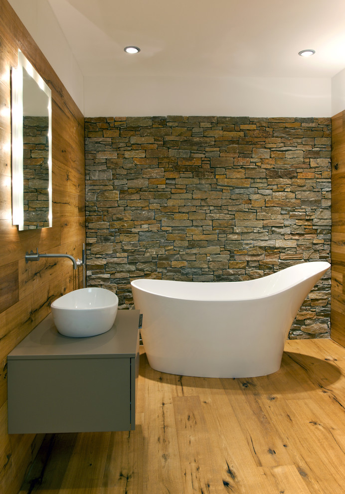 Example of a mid-sized trendy master medium tone wood floor freestanding bathtub design in Other with a vessel sink, flat-panel cabinets, gray cabinets and brown walls