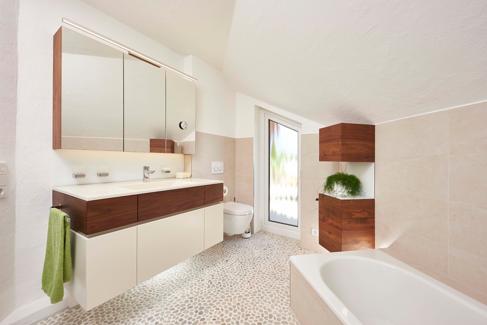 Mittelgroßes Modernes Duschbad mit flächenbündigen Schrankfronten, Einbaubadewanne, beigen Fliesen, weißer Wandfarbe, Kiesel-Bodenfliesen, integriertem Waschbecken, beigem Boden, weißen Schränken, Wandtoilette und weißer Waschtischplatte in Stuttgart