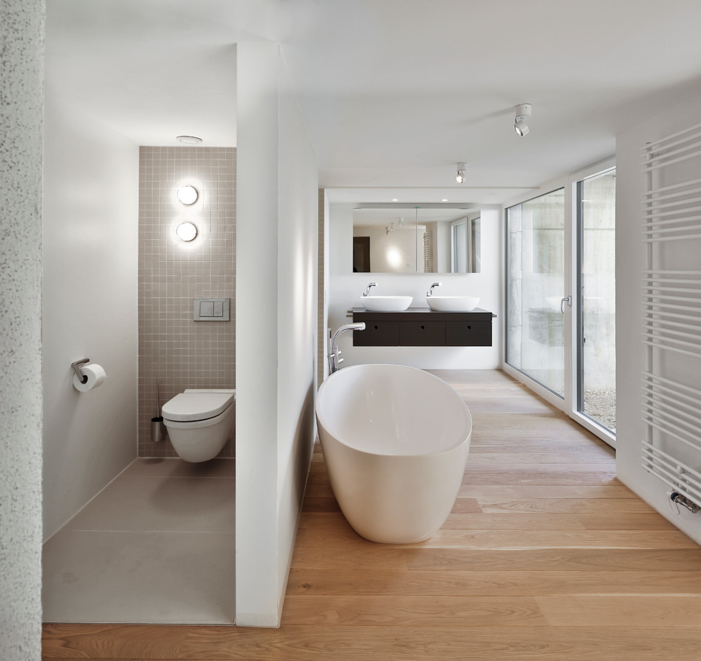 Modern inredning av ett brun brunt badrum, med ett fristående badkar, en vägghängd toalettstol, grå kakel, vita väggar, ett fristående handfat, släta luckor, svarta skåp, en dusch i en alkov, ljust trägolv, träbänkskiva och mosaik