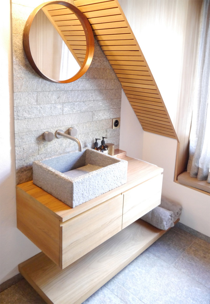 Bild på ett funkis badrum, med ett fristående handfat, släta luckor, skåp i ljust trä, träbänkskiva, grå kakel, cementkakel, vita väggar och skiffergolv