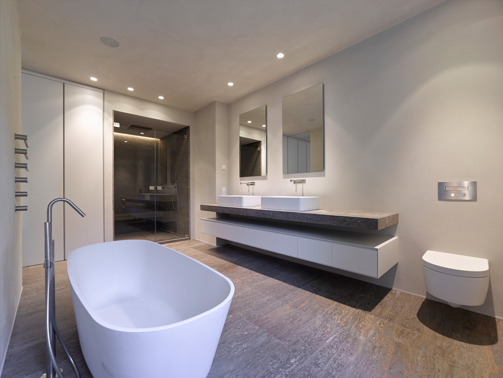 Design ideas for a large contemporary ensuite bathroom in Stuttgart with flat-panel cabinets, white cabinets, a freestanding bath, a built-in shower, a wall mounted toilet, beige tiles, beige walls, vinyl flooring, a vessel sink, granite worktops, brown floors and a hinged door.
