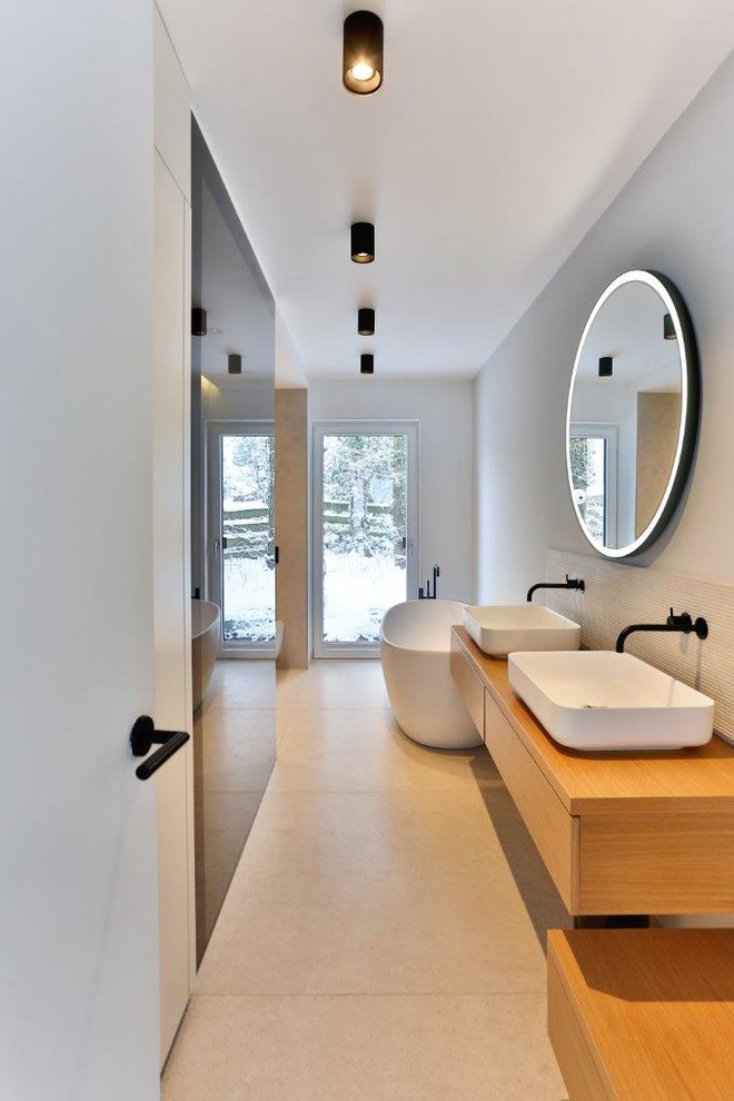 Свежая идея для дизайна: узкая и длинная ванная комната в стиле модернизм с плоскими фасадами, светлыми деревянными фасадами, отдельно стоящей ванной, душем без бортиков, инсталляцией, бежевой плиткой, керамогранитной плиткой, белыми стенами, полом из керамогранита, душевой кабиной, настольной раковиной, бежевым полом, открытым душем, тумбой под две раковины, подвесной тумбой, потолком с обоями и обоями на стенах - отличное фото интерьера