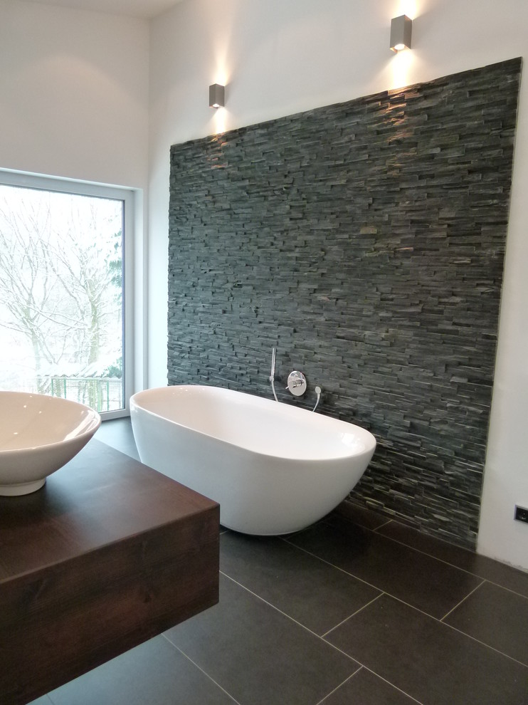 Cette image montre une salle de bain principale design de taille moyenne avec une baignoire indépendante, un carrelage gris, un carrelage noir, du carrelage en ardoise, un mur blanc, carreaux de ciment au sol, une vasque, un plan de toilette en bois et un sol marron.