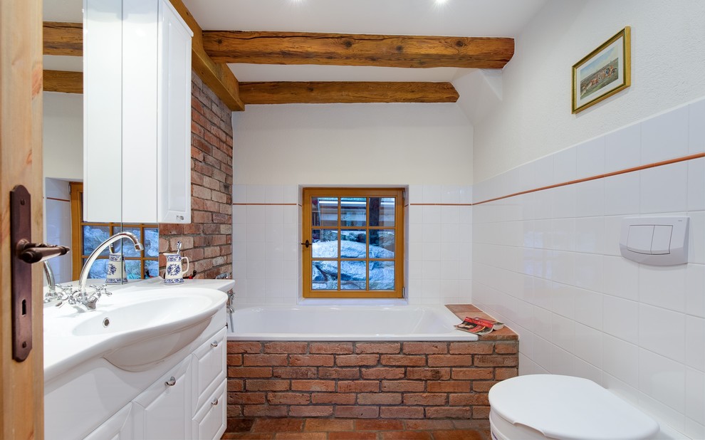 Inredning av ett rustikt litet badrum, med ett platsbyggt badkar, en vägghängd toalettstol, ett undermonterad handfat, klinkergolv i terrakotta, luckor med upphöjd panel, vita skåp, vit kakel och vita väggar