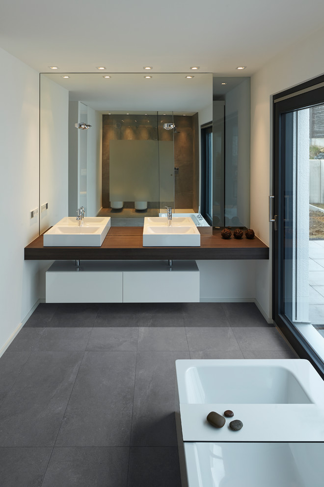 Cette image montre une grande salle de bain minimaliste avec un plan de toilette en bois, une baignoire indépendante, un mur blanc, un sol en ardoise, un placard à porte plane, des portes de placard blanches et une vasque.