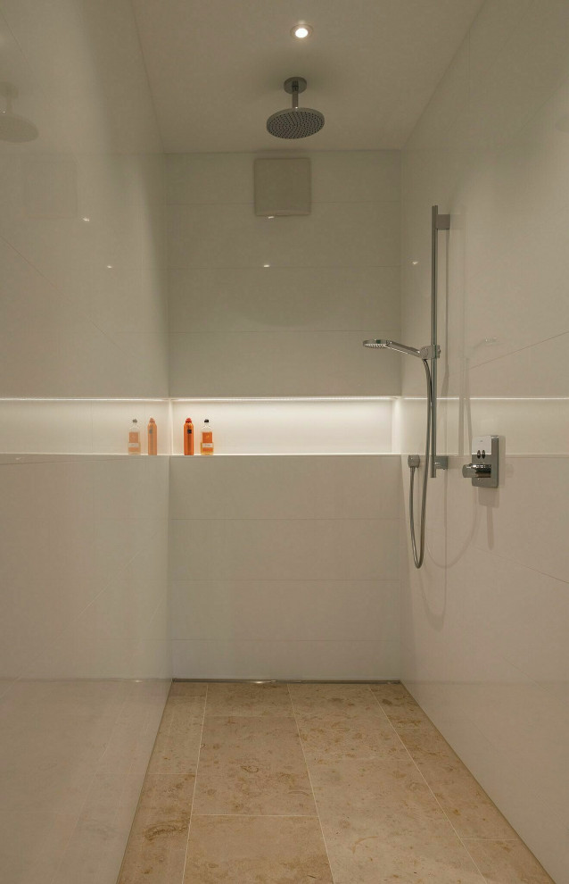 Стильный дизайн: большая, узкая и длинная главная ванная комната в стиле модернизм с белыми фасадами, отдельно стоящей ванной, открытым душем, раздельным унитазом, белыми стенами, монолитной раковиной, стеклянной столешницей, бежевым полом, открытым душем, белой столешницей, белой плиткой, плоскими фасадами, керамической плиткой, полом из известняка, тумбой под одну раковину, подвесной тумбой, многоуровневым потолком и панелями на стенах - последний тренд