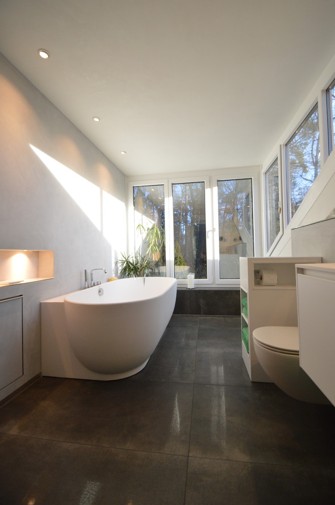 Modelo de cuarto de baño principal contemporáneo de tamaño medio con armarios con paneles lisos, puertas de armario blancas, bañera exenta, sanitario de pared, paredes grises y suelo negro