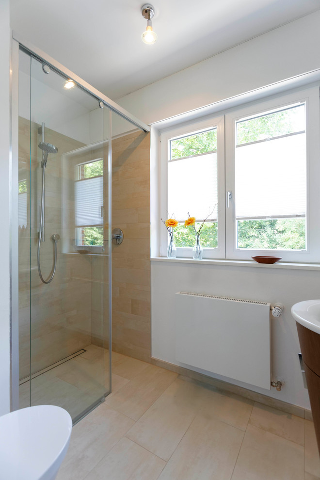 Modern inredning av ett badrum med dusch, med släta luckor, vita skåp, en kantlös dusch, en vägghängd toalettstol, beige kakel, keramikplattor, vita väggar, klinkergolv i keramik, ett avlångt handfat, träbänkskiva, beiget golv och dusch med skjutdörr