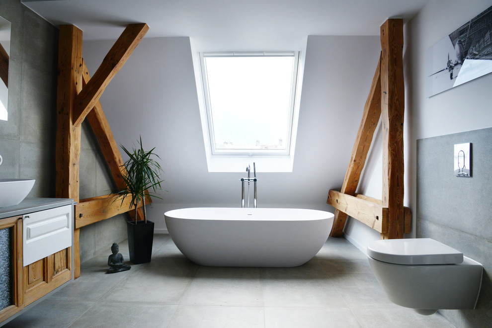 Aménagement d'une très grande salle de bain contemporaine avec une baignoire indépendante, WC suspendus, un carrelage gris, un mur blanc, sol en béton ciré, une vasque, un plan de toilette en bois et un placard avec porte à panneau surélevé.